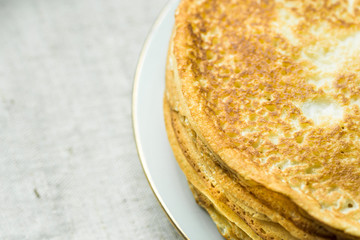 Stack of delicious golden crepes on white plate, on linen cloth background, top view