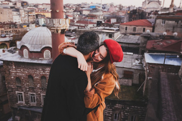 Couple in love hugging on roof with view of evening city at background. Casual style, autumn look, red beret and beige coat