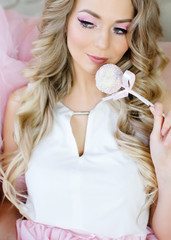 Nice blonde girl with clean skin and long hair, professional make-up, eats a cake pop. Puppet make-up, pink color, large portrait