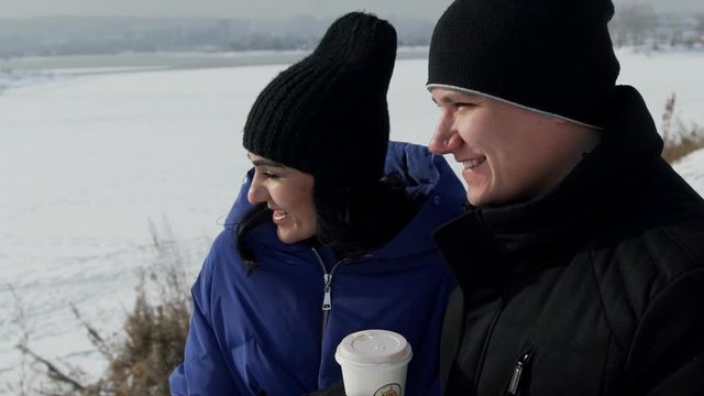 Man and woman drink coffee in picturesque place outdoors. Adult female with long dark hair, long eyelashes, black knitted cap, blue jacket with hood walk with caucasian man in down-jacket. They hold