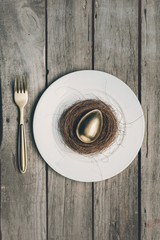 Top view of golden Easter egg in nest on white plate and fork on wooden table, Happy Easter concept