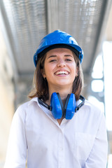 Vertical portrait of beautiful smiling engineer with blue hardhat and protective headphones around her neck against scaffolding. Selective focus