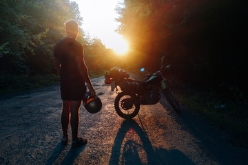 Young man holding helmet next to motorcycle scrambler loaded with camping gear for a moto tour staring into the sunset.