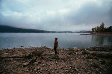 Fototapeta na wymiar Petite hipster girl standing near a foggy lake in the mountains surrounded by rocks and dead fallen trees dark cold adventure