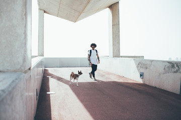 Young trendy hipster with tattoos crazy curly hair with his best friend dog love overlooking city white light wearing sunglasses and backpack walk on the urban bridge