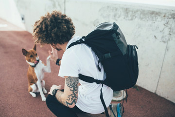 Young trendy hipster with tattoos crazy curly hair with his best friend basenji dog puppy give each...
