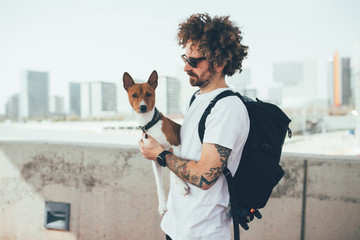 Young trendy hipster with tattoos crazy curly hair with his best friend dog love overlooking city white light wearing sunglasses and backpack