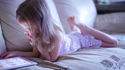 Little cute girl sitting on sofa and playing with tablet