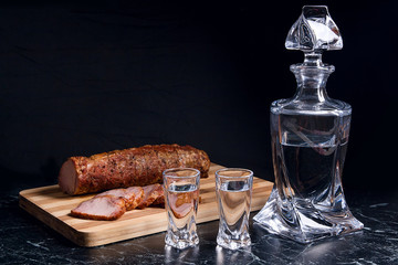Bottle and shot glasses with vodka with slices of smoked meat on wooden cutting board.