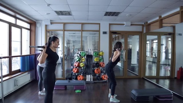 Trainer do exercises with barbell with her group of women in studio. Young slim woman in black training suit, grey snickers, with flowing disheveled dark hair with bar on her shoulders stand in front