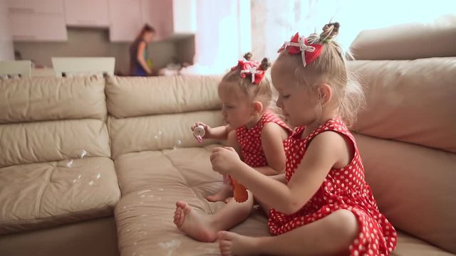 Two little twin sisters in charming red dresses are blowing soap bubbles cheerfully while sitting in the room barefoot.