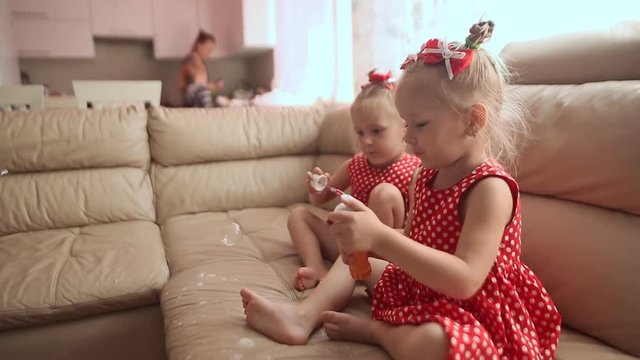 Two little twin sisters in charming red dresses are blowing soap bubbles cheerfully while sitting in the room barefoot.