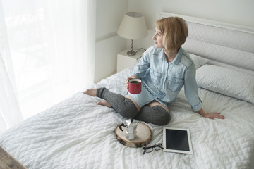 Girl with cup in her hand, kettle and tablet on a white bed looking in window