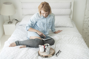 Girl drinking morning coffee on a white bed working on tablet in high stockings