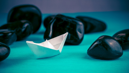 Lonely White Paper Ships between Abstract Black Rock Stones on Blue Background. Blue-Violet Colours