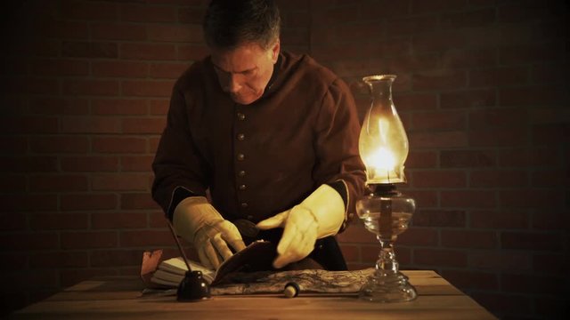 In a room lit only by the light from an oil lamp a mature somber civil war officer using a dip pen writes in a journal.
