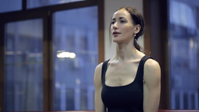 Brunette sportswoman do exercise with orange dumbbells in gym. Young slim woman in black training suit, small earnings, natural make raise hands with weight in front of her. They are straight. Every