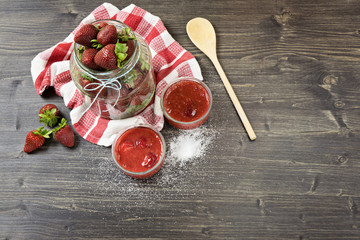 Large jar with fresh strawberries and bowls with strawberry jam on the wooden background, top view