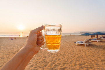 Sunset over sandy beach and one glass of cold beer in hand of happy tourist.