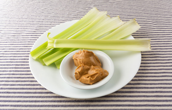 Celery stalks with peanut butter on a white plate atop a blue striped tablecloth.
