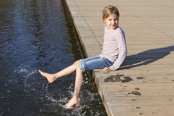 Cute little girl sitting on a wooden platform by the river or lake while splashing water by feet on warm summer day
