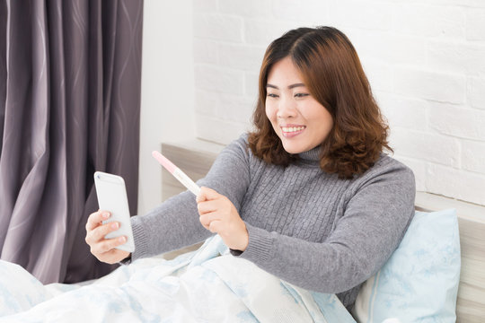 Happy asian woman holding pregnancy test and taking selfie by smartphone