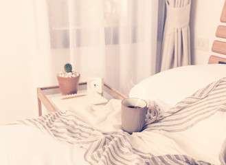 Cup of coffee on cozy white bed with cactus flowerpot,memo pat and alarm clock on wood bed side table