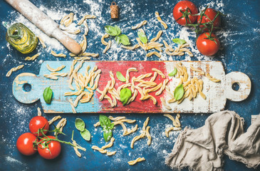 Ingredients for cooking Italian dinner. Fresh pasta casarecce, cherry-tomatoes, basil leaves and bottle of olive oil on colorful wooden board over dark blue background. Top view, horizontal
