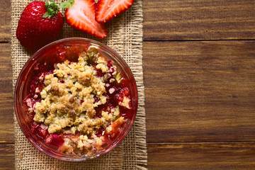 Baked strawberry and rhubarb crumble in glass bowl, photographed overhead on dark wood with natural light