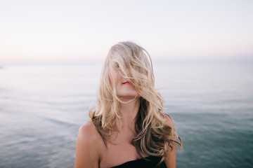 Beautiful young blonde in black dress walking along the seashore and posing at sunrise in summer