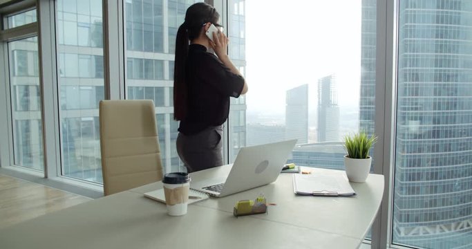 Asian girl in a modern office talking on the phone standing desktop, view the skyscrapers outside the window