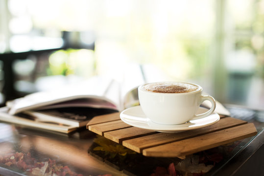 Cup of cappuccino with magazine on the table, coffee shop background, bright tone