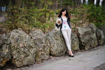 Brunette gypsy girl with yorkshire terrier dog posed against stones on park. Model wear on leather jacket and t-shirt with ornament, pants and shoes with high heels.