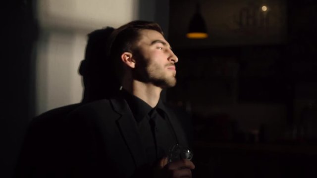 The serious man with black hair and suit is drinking light beer and standing near the wall of the dack pub. The close-up side portrait.