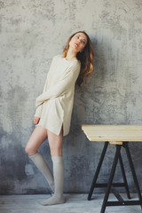 young woman in oversize knitted sweater posing by the grey wall in modern apartment. Casual lifestyle at home