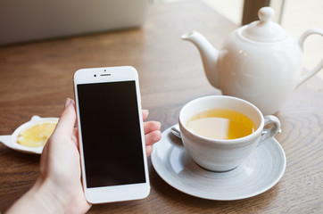 Cropped view of female hand holding smart phone with blank screen for your text. Woman reading messages on cell phone with cafe interior background while drinking hot cup of tea with lemon