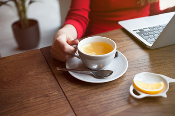Cropped view of female hand holding smart phone with blank screen for your text. Woman reading messages on cell phone with cafe interior background while drinking hot cup of tea with lemon