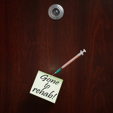 Gone to rehab! The text is written on a paper note pinned to a wooden door with a steel needle attached to a syringe. A peephole is visible. Concept photograph.