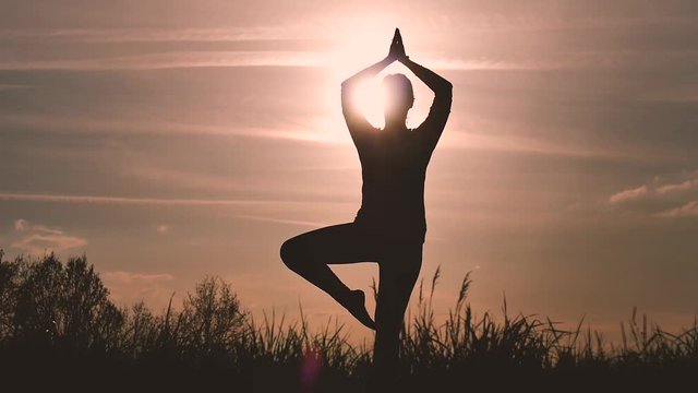 silhouette of a girl does some yoga poses in backlight during sunset in the countryside to train and take care of herself, tree pose and meditation