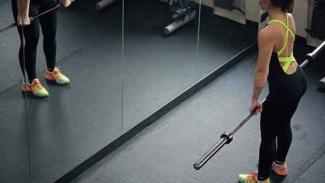 Young beautiful woman doing deadlift in front of mirror in gym. Attractive redheaded sportswoman in black training suit with green parts on back, colorful snickers with funny patterns, with tattoo on