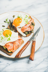 Healthy breakfast sandwiches. Salmon, avocado, fried egg, sauted green beans and fresh sprouts sandwiches in white plate over marble background, selective focus. Clean eating, weight loss food concept