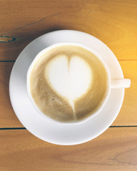 A white cup of cappuccino with heart shape foam on top on a wooden table background
