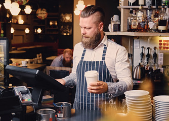 The bearded coffee seller holds a coffee cup at the counter in a coffee shop.