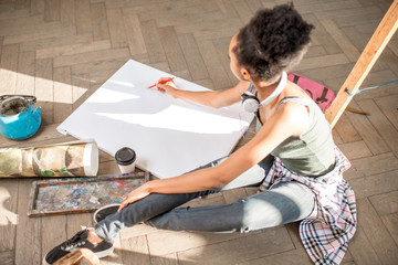 Female student drawing on the white canvas sitting on the floor. View from above