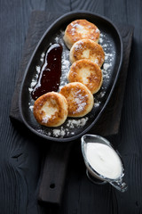 Cheesecakes topped with powdered sugar and served with jam and sour cream, black wooden background, studio shot