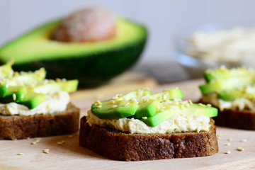 Hummus avocado sandwiches on a wooden board, avocado half, hummus in a glass bowl. Veggie sandwiches made with rye bread, avocado slices, hummus and roasted sesame seeds. Closeup