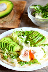 Poached egg, avocado slices, salad mix, sauce, spices in a flour tortilla. Avocado half on a wooden board, salad mix in a glass bowl on a table. Healthy and diet breakfast idea. Vertical photo