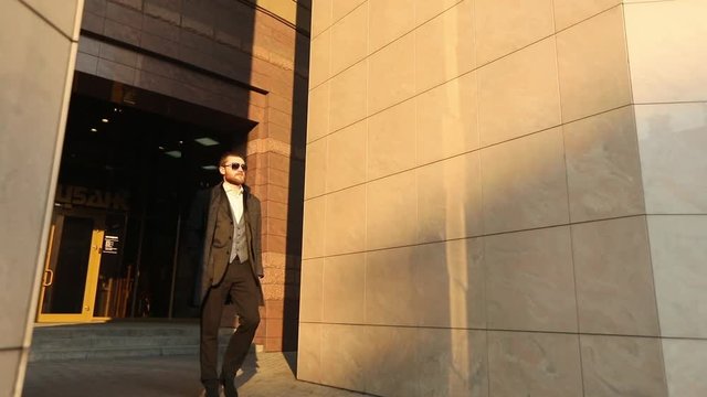 The fashionably dressed man in the dack suit, coat and sunglasses is keeping his hand in the pocket and leaving the building.