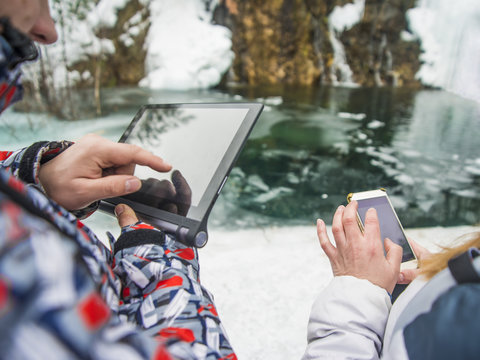 Back View Of Man And Tourist Woman Using Laptop And Mobile Phone With Gps Navigation On Winter Lake And Snowy Mountain Background. No Face. Unrecognizable Person