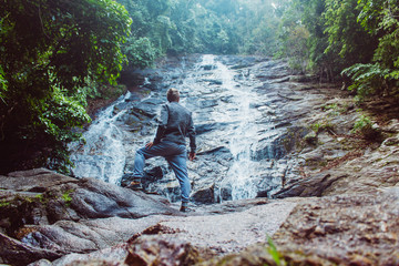 Obraz premium Male posing against waterfall . Back view of young man standing on rock near beautiful water stream flowing down stiff slope.